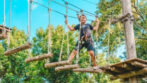 Boy walking on suspended logs under a blue sky with trees in the background